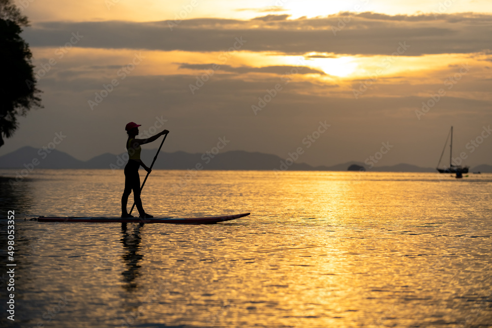 Confidence mature Asian woman sitting on paddle board and paddleboarding passing beautiful sea at summer sunset. Healthy female enjoy outdoor active lifestyle and water sports on holiday vacation