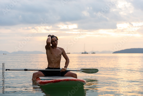 Confidence young Asian man sitting on paddle board and paddleboarding passing beautiful sea at summer sunset. Healthy strong male enjoy outdoor active lifestyle and water sports on holiday vacation © CandyRetriever 