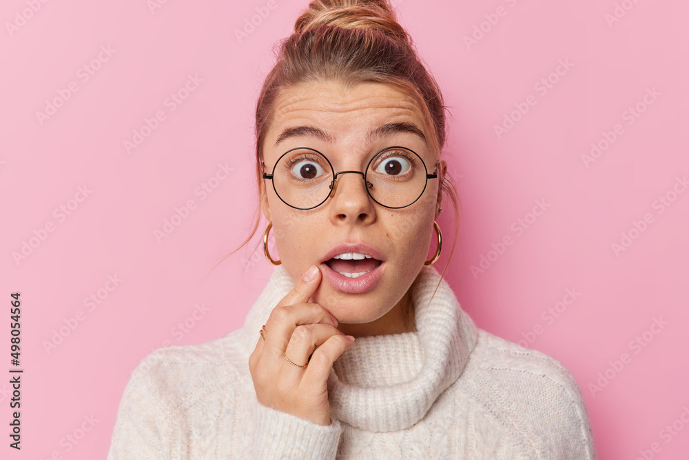 Scared woman stares shocked holds breath has mouth opened from amazement wears spectacles and sweater with collar reacts on something breathtaking isolated over pink background. Human reaction Scared woman stares shocked holds breath has mouth opened from amazement wears spectacles and sweater with collar reacts on something breathtaking isolated over pink background. Human reaction