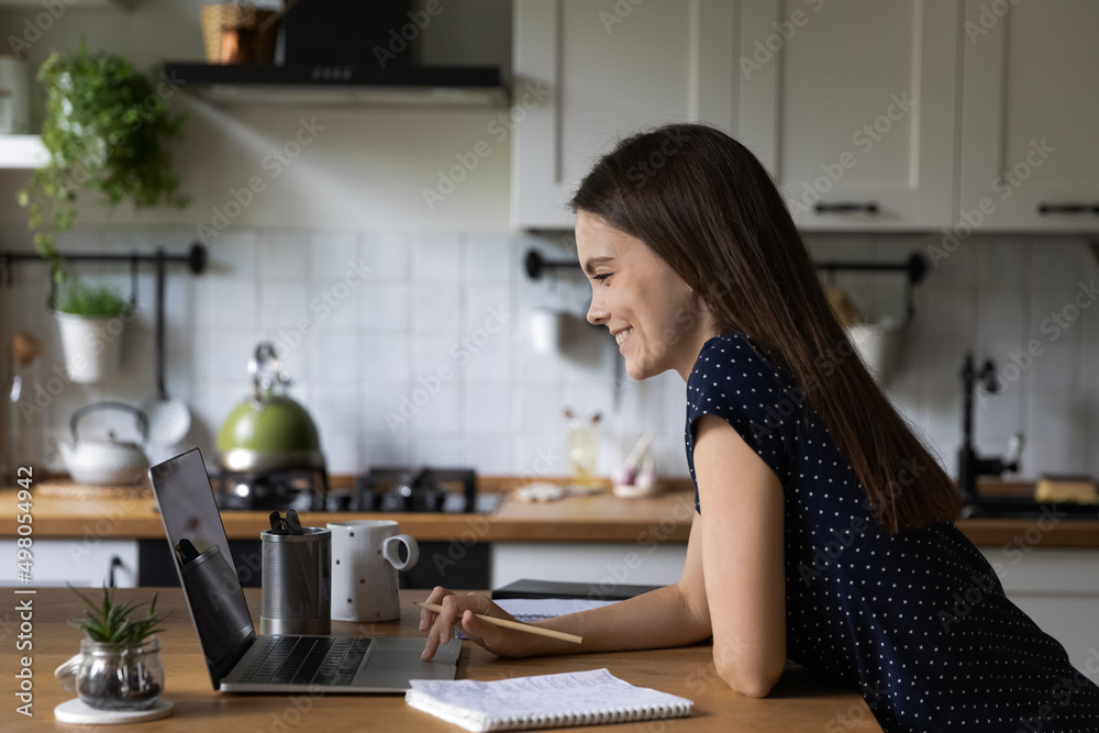 Young woman studying or working on laptop standing lean at kitchen table looks at screen, read information, web surfing, e-learns, gain new knowledge. Webinar, learning at home use modern tech concept Young woman studying or working on laptop standing lean at kitchen table looks at screen, read information, web surfing, e-learns, gain new knowledge. Webinar, learning at home use modern tech concept