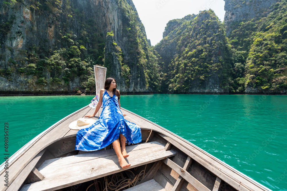 Young beautiful Asian woman in blue dress sitting on the boat passing island beach lagoon in summer sunny day. Happy female relax and enjoy outdoor lifestyle together on summer vacation in Thailand