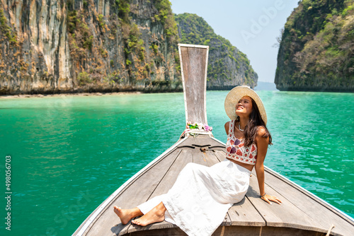 Young beautiful Asian woman in blue dress sitting on the boat passing island beach lagoon in summer sunny day. Happy female relax and enjoy outdoor lifestyle together on summer vacation in Thailand
