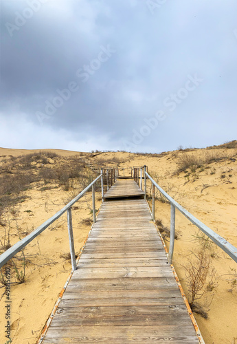 The staircase rises to a sandy dune. Climbing to the top in the desert. Sarykum dune. Dagestan  Russia. A unique sandy mountain in the Caucasus on a cloudy day. Grass grows on a sand dune.