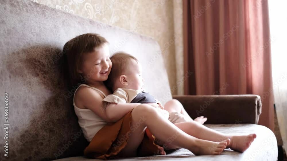 Funny children siblings play together on the sofa. Toddler and baby in a reusable diaper at home in a real interior. Sibling relationships. Sister combing and hugging little brother