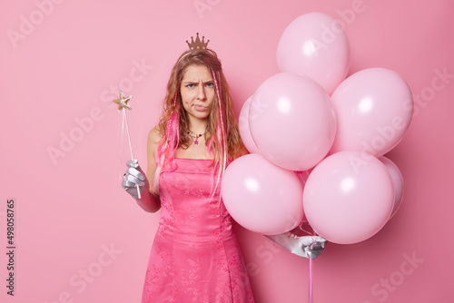Annoyed displeased long haired young woman wears crown dress and gloves holds magic wand and bunch of inflated balloons being irritated celebrates something on party isolated on pink background