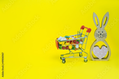 Wooden Easter Bunny and a shopping cart with candies on a yellow background. The concept of shopping, gifts, holiday, invitation, gratitude, congratulation, postcard . High quality photo