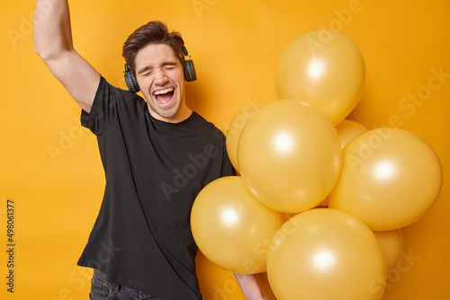 Cheerful adult man exclaims from joy keeps arm raised up sings favorite song dressed in casual black t shirt wears stereo headphones on ears holds bunch of inflated balloons isolated over yellow wall © wayhome.studio 