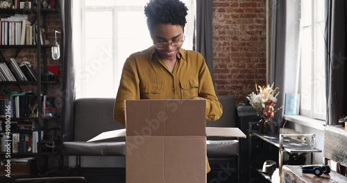 Curious smiling Black gen z female unpack postal parcel open big box with consumer goods purchased on sale at ecommerce web store. Happy millennial female client receive unbox mail delivery package