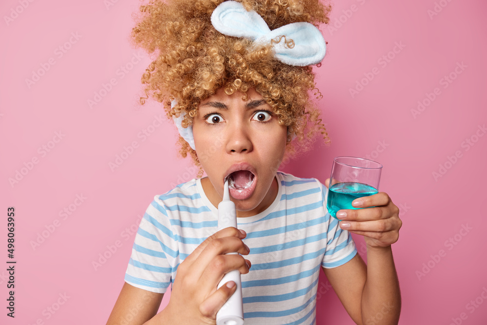 Shocked young woman with curly hair wears headband and casual striped t shirt brushes teeth with electric brush holds glass of blue mouthwash isolated over pink background. Daily hygiene concept Shocked young woman with curly hair wears headband and casual striped t shirt brushes teeth with electric brush holds glass of blue mouthwash isolated over pink background. Daily hygiene concept