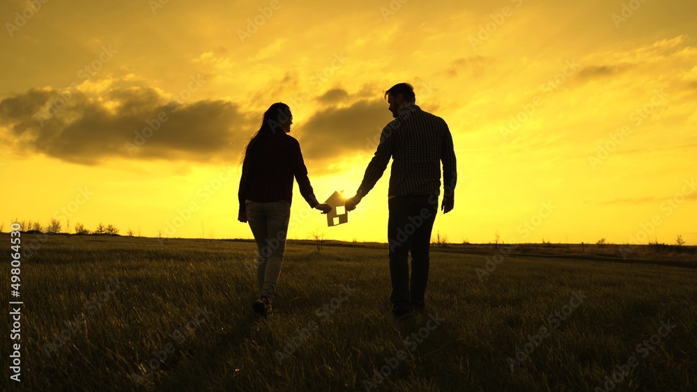 Family's hands are holding paper house at sunset, sun is shining through window. Symbol of house, happiness. Concept of building house for family. Dream to buy house. Family silhouette, family home