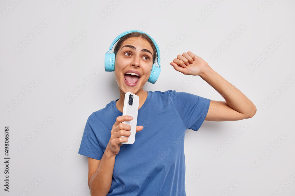 Beautiful young European woman dances and sing song holds modern smartphone as if microphone uses wireless stereo headphones wears casual t shirt has upbeat mood isolated over white background Beautiful young European woman dances and sing song holds modern smartphone as if microphone uses wireless stereo headphones wears casual t shirt has upbeat mood isolated over white background