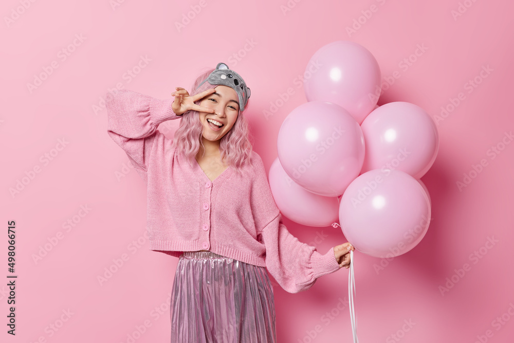 Happy upbeat Asian woman makes peace gesture over eye smiles gladfully poses with inflated balloons dressed in festive clothes celebrates special occasion on costume party isolated on pink wall Happy upbeat Asian woman makes peace gesture over eye smiles gladfully poses with inflated balloons dressed in festive clothes celebrates special occasion on costume party isolated on pink wall