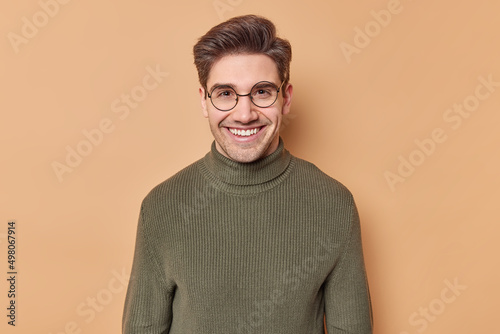 Portrait of positive guy with dark hair smiles happily wears round transparent eyeglasses and sweater with collar hears something funny isolated over brown background. Happy emotions concept © wayhome.studio 