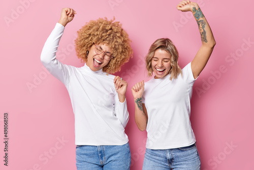 Horizontal shot of cheerful upbeat women dance carefree dressed in white t shirts and jeans move with rhythm of music isolated over pink background foolish around. Dance like nobody watching