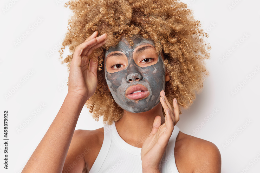 Photo of serious charming curly woman touches face gently applies clay facial mask to look beautiful wears casual t shirt poses against white background shows bare shoulders. Skin care concept Photo of serious charming curly woman touches face gently applies clay facial mask to look beautiful wears casual t shirt poses against white background shows bare shoulders. Skin care concept