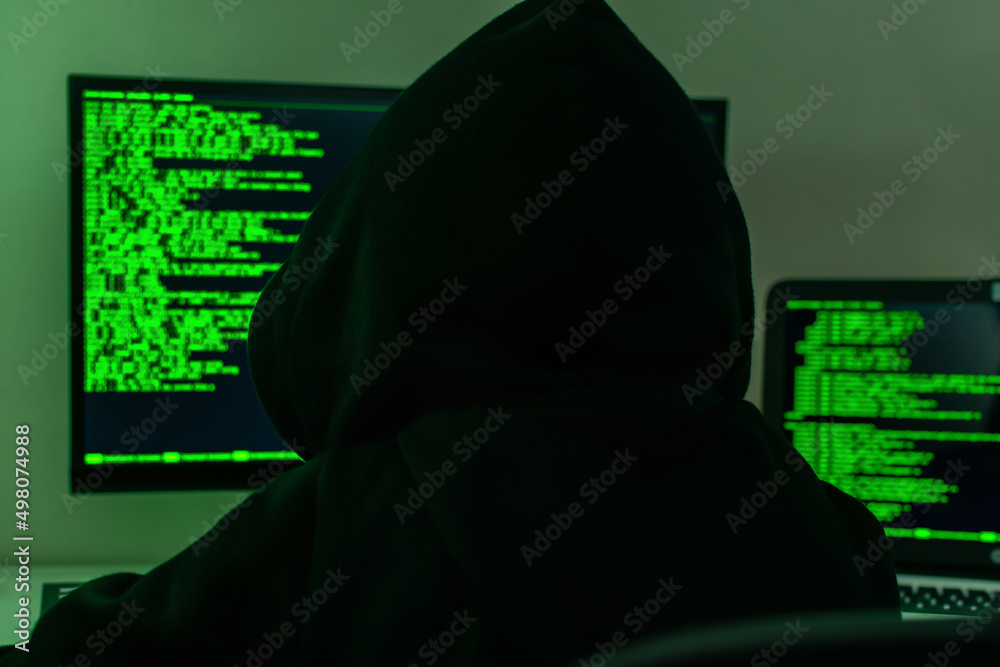 Rear view a hacker in a black hood is sitting in front of a computer monitor and writing a security hacking code in a dark room. The concept of information protection, cyber security, cybercrime. Rear view a hacker in a black hood is sitting in front of a computer monitor and writing a security hacking code in a dark room. The concept of information protection, cyber security, cybercrime.