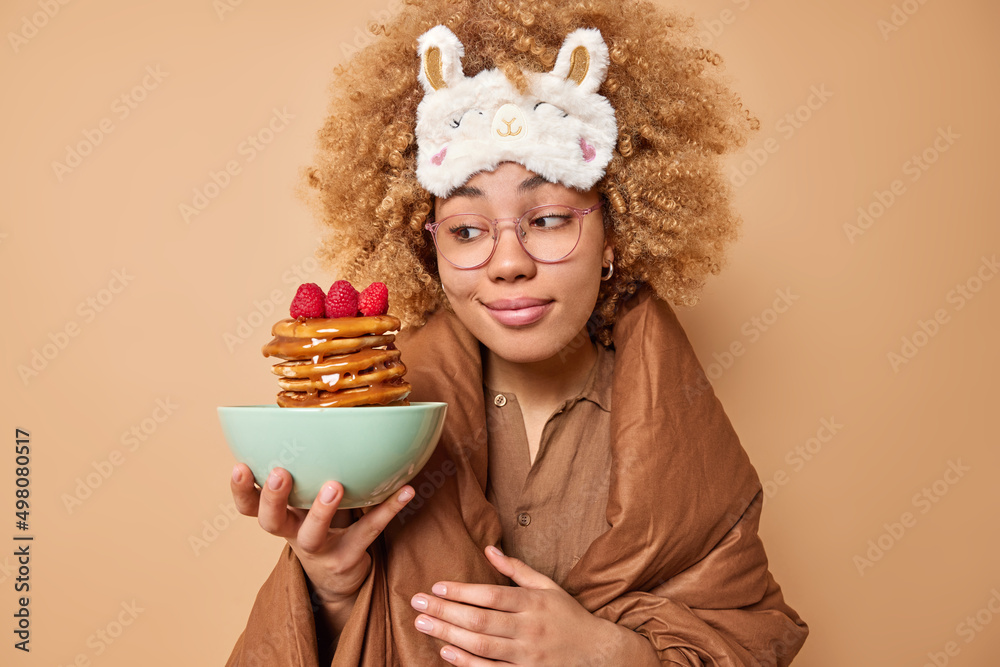 Lovely young beautiful woman with curly bushy hair wears sleepmask looks at appetizing pancakes with syrup and raspberries wrapped in duvet has delicious breakfast isolated over beige background. Lovely young beautiful woman with curly bushy hair wears sleepmask looks at appetizing pancakes with syrup and raspberries wrapped in duvet has delicious breakfast isolated over beige background.
