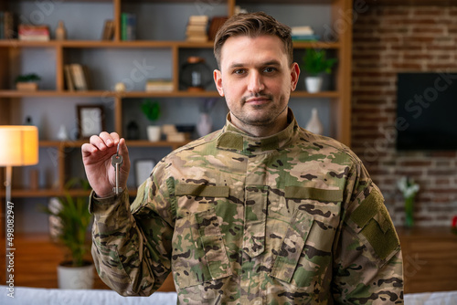 Indoor shot of military man wearing camouflage uniform and hat holding house keys, mortgage assistance from a veterans organization, extremely happy soldier male.