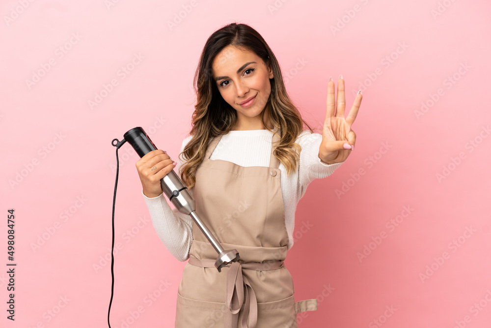 Young woman using hand blender over isolated pink background happy and counting three with fingers Young woman using hand blender over isolated pink background happy and counting three with fingers