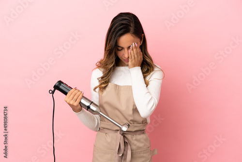 Young woman using hand blender over isolated pink background with tired and sick expression
