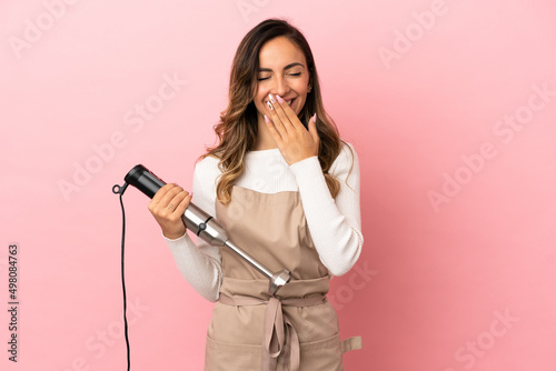 Young woman using hand blender over isolated pink background happy and smiling covering mouth with hand