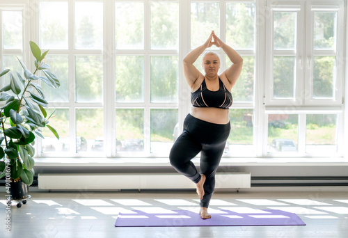 Plump  chubby woman doing yoga standing in a tree pose. Female keeps balance by doing fitness while standing on a sports mat. Room with large high windows to the floor, letting in a lot of sunlight