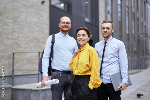 Confident young confident business colleagues standing smiling outside office building looking forward with blueprints and laptop. Team of economists or real estate managers in casual clothes outdoors