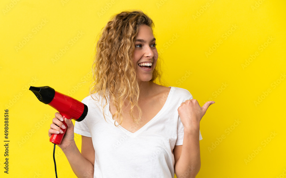 Young blonde woman holding a hairdryer isolated on yellow background pointing to the side to present a product Young blonde woman holding a hairdryer isolated on yellow background pointing to the side to present a product