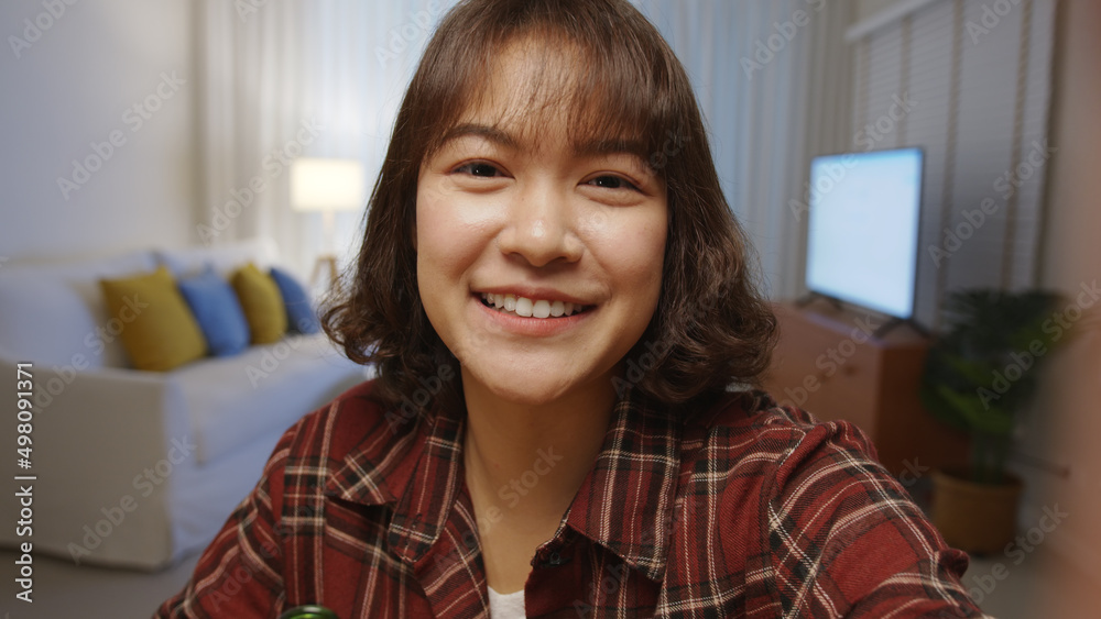 Fototapeta premium Selfie view of young happy attractive asia woman enjoy relax night party event online celebration festive with friends at home clinking beer cheers with bottle toasting drinking via online video call.