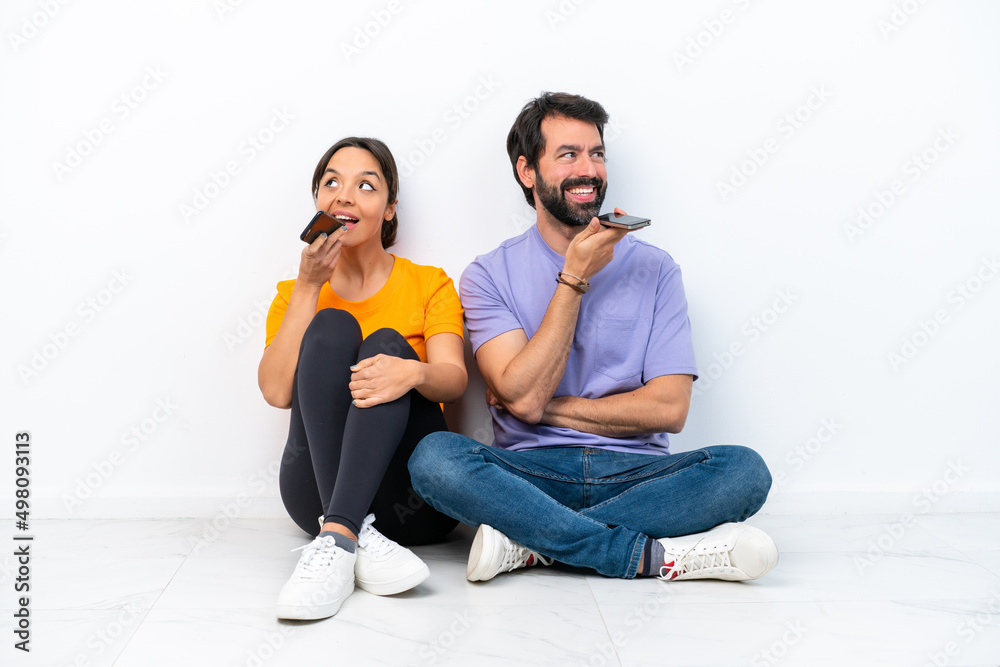 Young caucasian couple sitting on the floor isolated on white background sending a message voice or email with the mobile Young caucasian couple sitting on the floor isolated on white background sending a message voice or email with the mobile