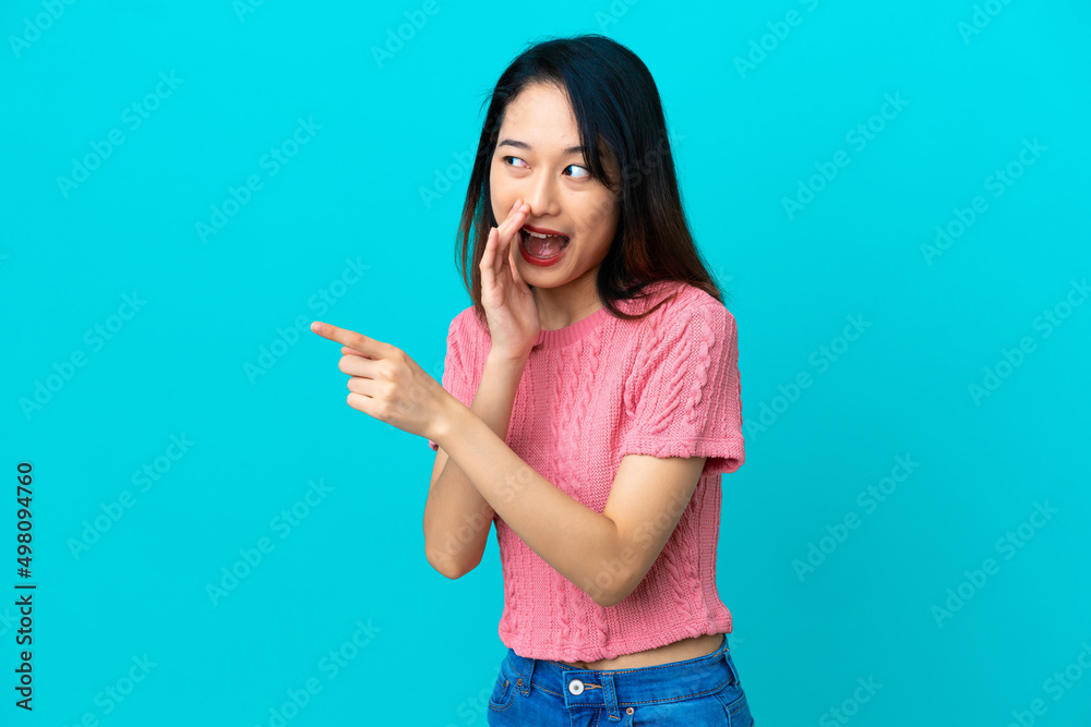 Young Vietnamese woman isolated on blue background pointing to the side to present a product and whispering something Young Vietnamese woman isolated on blue background pointing to the side to present a product and whispering something