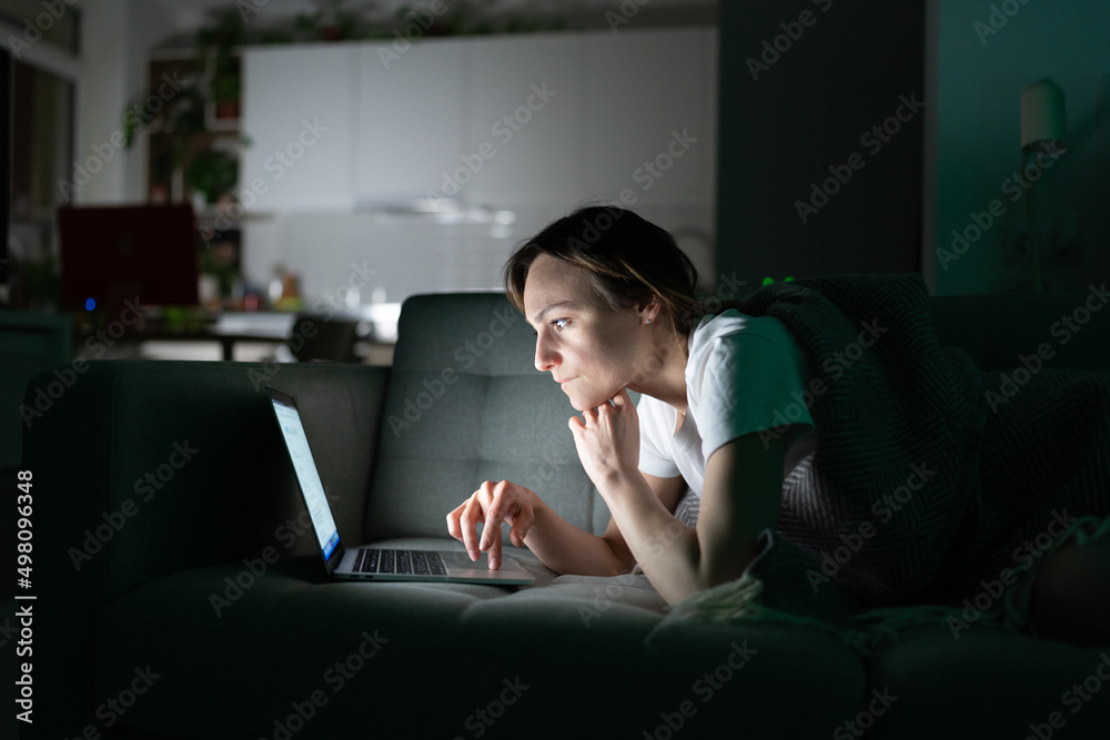 Focused young woman working online on laptop at home in the night, looking at screen, thinking, reading news media. Concentrated girl lying on sofa in dark room, using computer, suffers from insomnia Focused young woman working online on laptop at home in the night, looking at screen, thinking, reading news media. Concentrated girl lying on sofa in dark room, using computer, suffers from insomnia