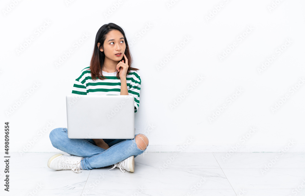 Young Vietnamese woman with a laptop sitting on the floor isolated on white wall thinking an idea while looking up Young Vietnamese woman with a laptop sitting on the floor isolated on white wall thinking an idea while looking up
