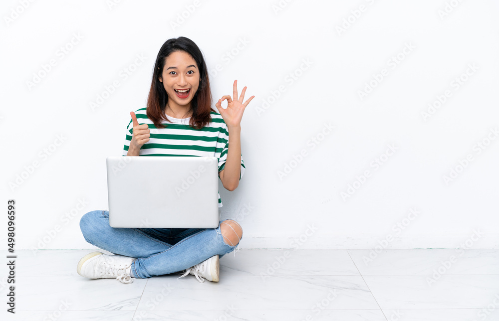 Young Vietnamese woman with a laptop sitting on the floor isolated on white wall showing ok sign and thumb up gesture Young Vietnamese woman with a laptop sitting on the floor isolated on white wall showing ok sign and thumb up gesture