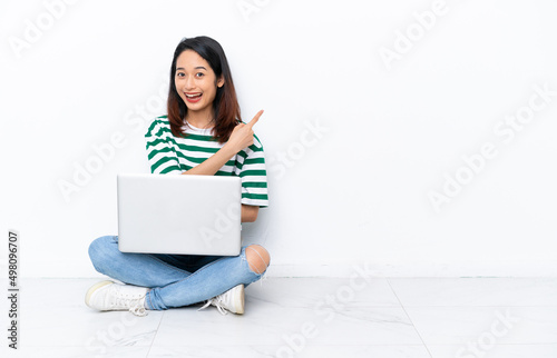 Young Vietnamese woman with a laptop sitting on the floor isolated on white wall surprised and pointing side