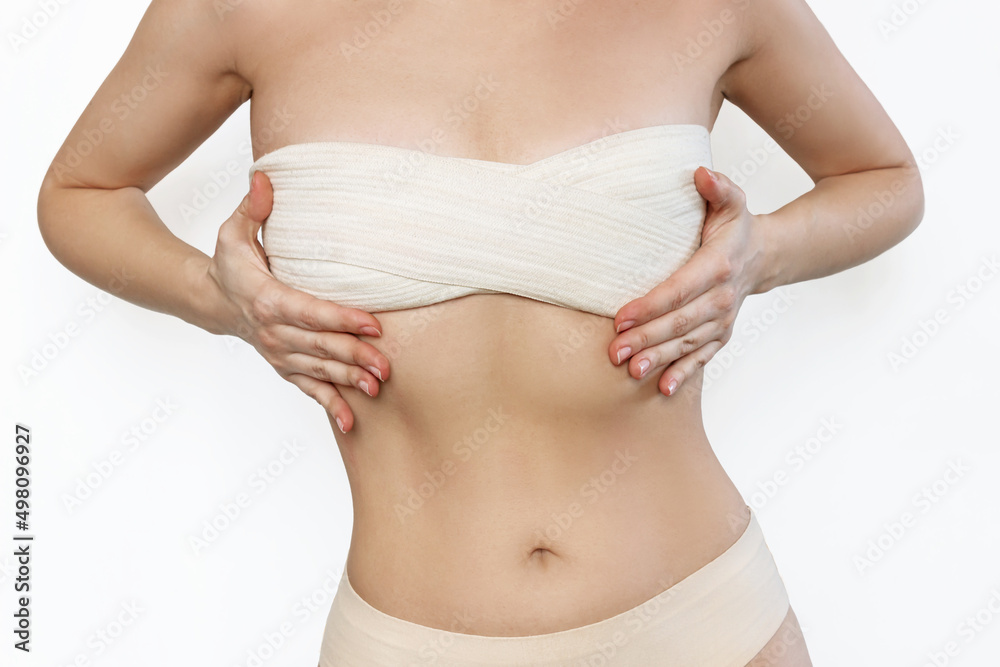Cropped shot of young slim woman wrapped in elastic bandage after breast augmentation isolated on white background. Result of lifting. Pain after plastic surgery. Girl is trying to hide her big breast