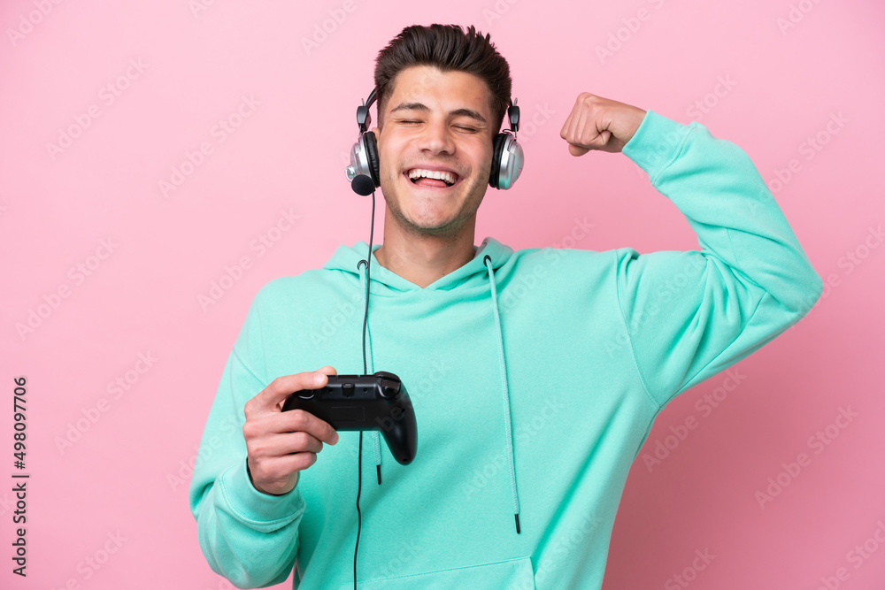 Young handsome caucasian man playing with a video game controller isolated on pink background doing strong gesture Young handsome caucasian man playing with a video game controller isolated on pink background doing strong gesture