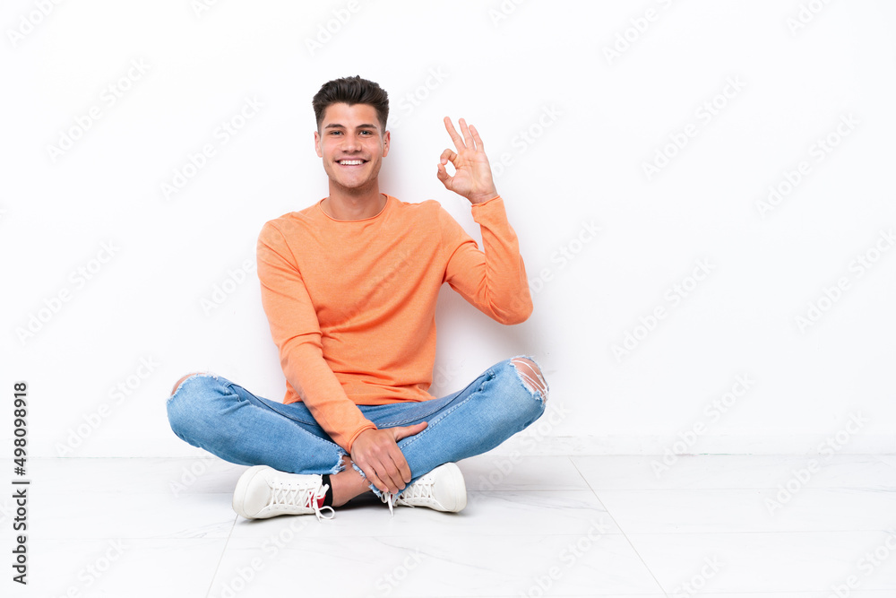 Young man sitting on the floor isolated on white background showing ok sign with fingers Young man sitting on the floor isolated on white background showing ok sign with fingers