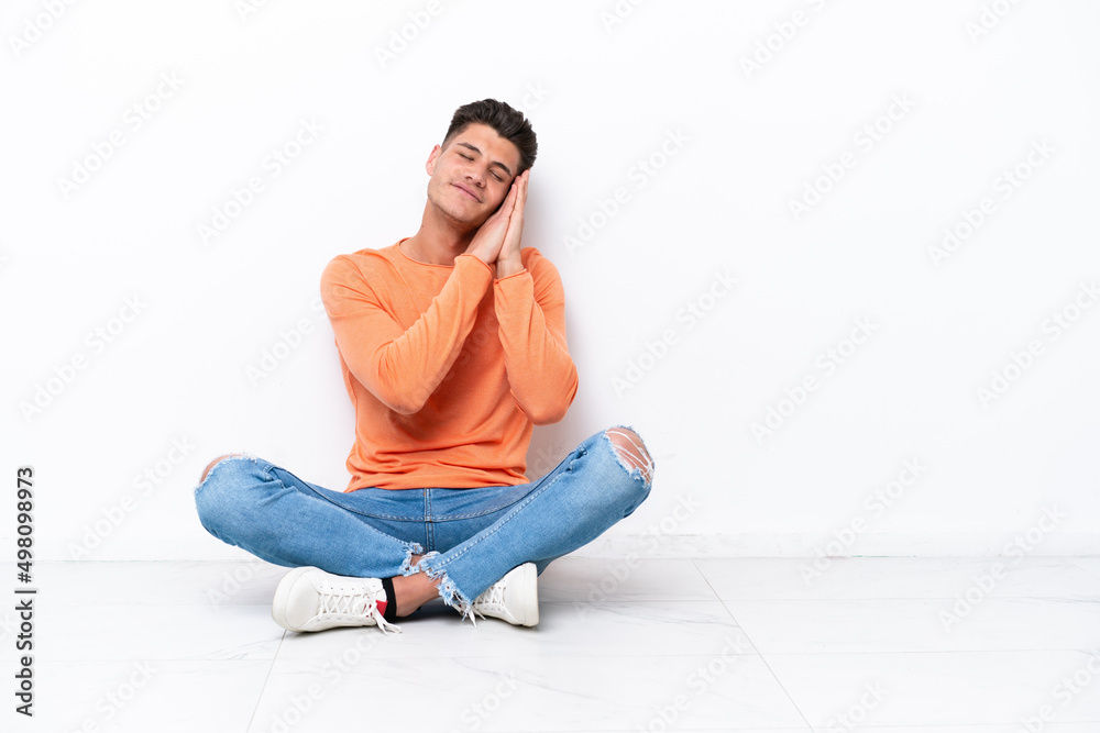 Young man sitting on the floor isolated on white background making sleep gesture in dorable expression Young man sitting on the floor isolated on white background making sleep gesture in dorable expression