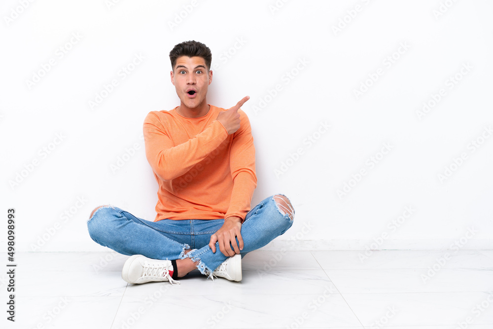Young man sitting on the floor isolated on white background surprised and pointing side Young man sitting on the floor isolated on white background surprised and pointing side