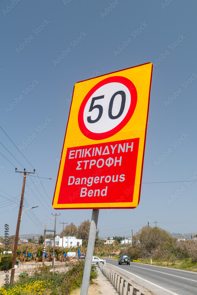 Dangerous bend warning and reduce speed sign. Reduce speed inscription in English and Greek languages. Dangerous bend warning and reduce speed sign. Reduce speed inscription in English and Greek languages.