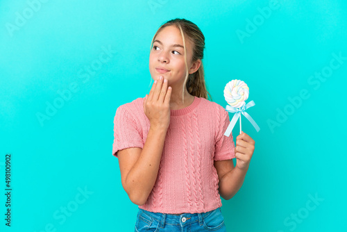 Little Caucasian girl holding a lollipop isolated on blue background looking up while smiling