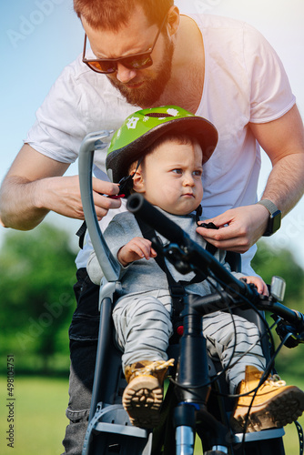 Caring father fastening helmet for his funny grumpy baby boy