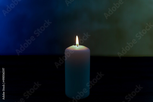 Biała świeca płonąca na ciemnym tle posiadającym delikatne niebieski i żółte zabarwienie. Hołd i wyraz solidarności z ofiarami wojny w Ukrainie. photo