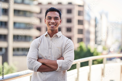 A young attractive Asian man stands in front of city buildings with arms crossed