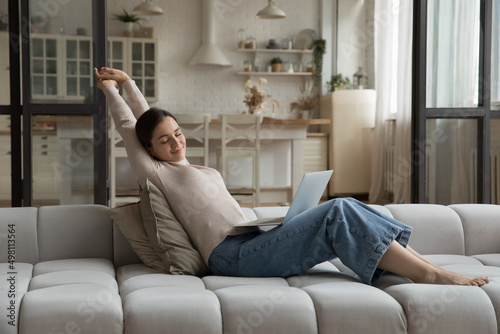 Happy satisfied millennial laptop user woman relaxing on comfortable spacious couch, stretching body with sleepy closed eyes, breathing fresh air, enjoying home leisure, feeling good, stress relief