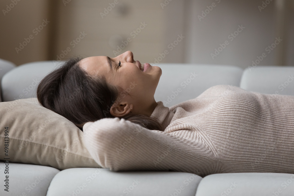 Happy cheerful millennial 20s girl resting on couch at home with closed eyes, smiling, laughing, dreaming, enjoying break, leisure time pause, breathing fresh air, lying on back. Side view Happy cheerful millennial 20s girl resting on couch at home with closed eyes, smiling, laughing, dreaming, enjoying break, leisure time pause, breathing fresh air, lying on back. Side view