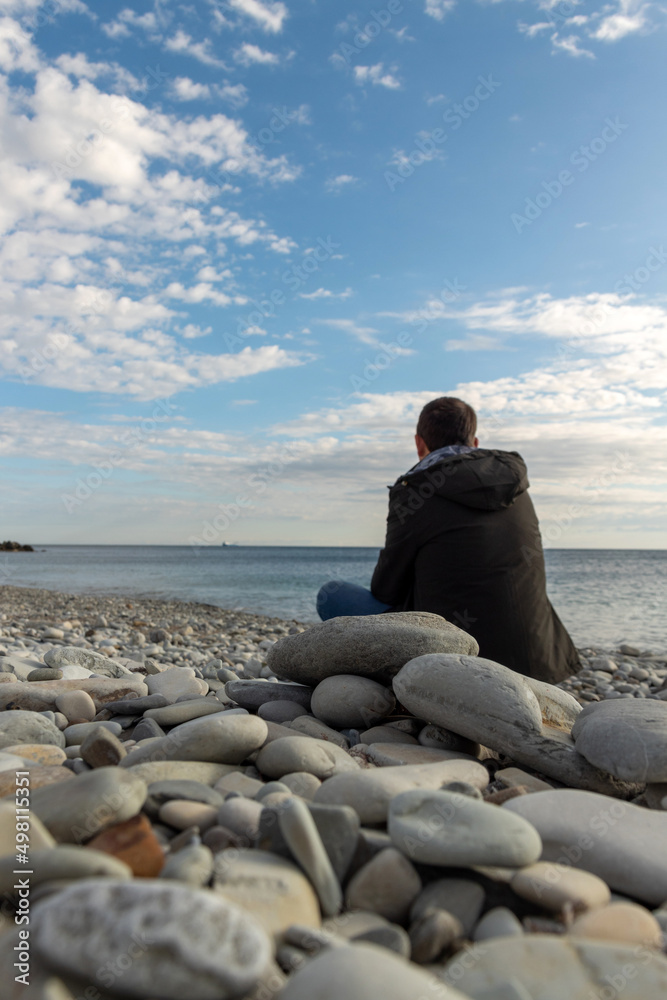 Focus on the rocks on the shore. Dark-haired man in the course is sitting on the shore, with his back in the frame and looking at the ship at sea. Selective focus. Picture about leisure of people. Focus on the rocks on the shore. Dark-haired man in the course is sitting on the shore, with his back in the frame and looking at the ship at sea. Selective focus. Picture about leisure of people.