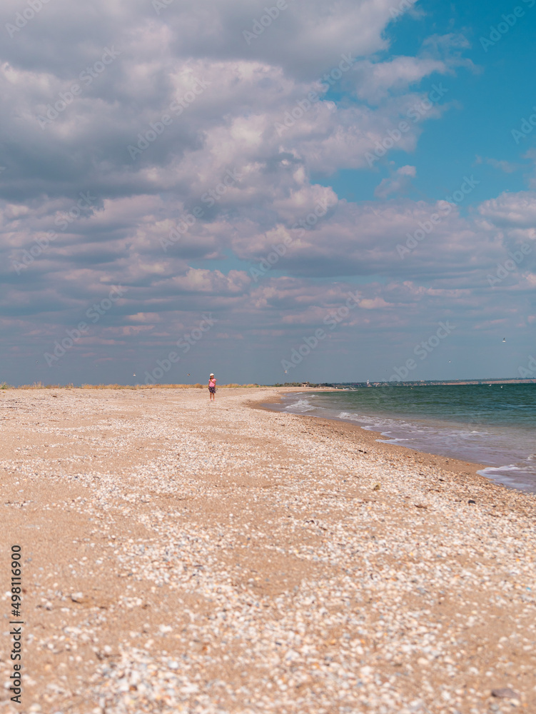 Obraz premium Happy child in a straw hat running jumping having fun on empty autumn sea beach. Blond girl walking on white sand road with blue cloudy sky background. Lifestyle photo Happiness freedom travel concept