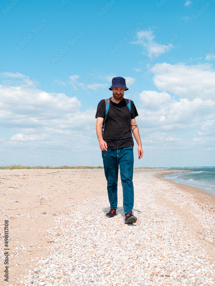 Smiling bearded millennial man in bucket hat black t-shirt walking on empty beach. Authentic male tourist lifestyle photo. Hipster guy outdoor. Solo travel adventure concept Active walking backpacking